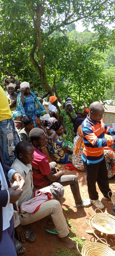 A group of people, predominantly women in colorful traditional clothing and head coverings, are seated outdoors in a shaded area. A few men are also present, one wearing an orange and blue striped shirt. Some people are seated on the ground, while others are elevated slightly. Woven baskets are visible in the foreground. A tree provides shade. The setting appears to be a gathering or meeting in a rural location.