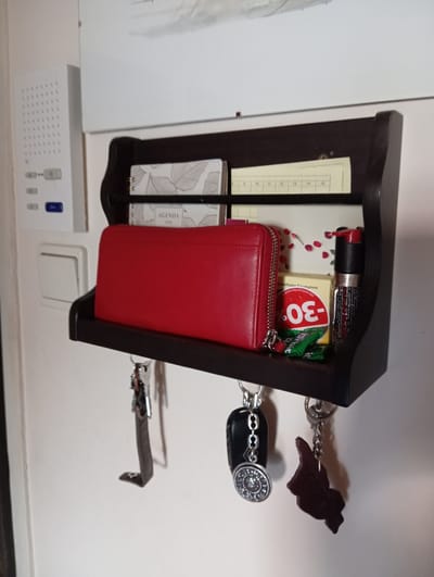 A dark wood shelf is mounted on a white wall. The shelf holds a red wallet, a small notebook, and a few other items. Below the shelf, keys with various charms are hanging from hooks. To the left of the shelf, a white intercom system is visible. The scene is indoors, likely in a hallway or entrance area. The objects are arranged in a tidy manner, suggesting a personal storage space.