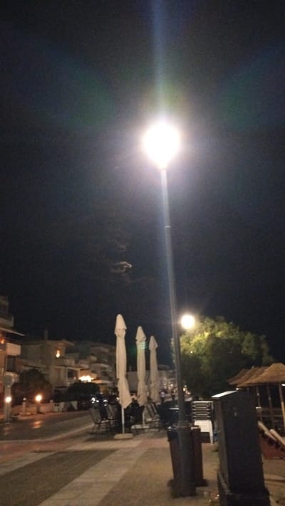 This is a night-time photograph taken in Perea, Greece. A tall street lamp with a bright light at its apex dominates the center of the frame. The sky above is dark, with a faint, swirling pattern visible, possibly clouds or smoke. To the left of the street lamp, there are buildings and trees. A sidewalk is visible in the foreground, with a street running alongside it. Several closed white umbrellas are positioned on the sidewalk, suggesting the area might be an outdoor seating area for a cafe or restaurant. Chairs and tables are also visible, some set up and some stacked. A person can be seen sitting at one of the tables. Further to the right, there are more trees and what appear to be thatched-roof structures, possibly part of a beach bar or similar establishment. The ground is paved, with visible lines or patterns. The overall impression is of a quiet street scene at night in a coastal town.