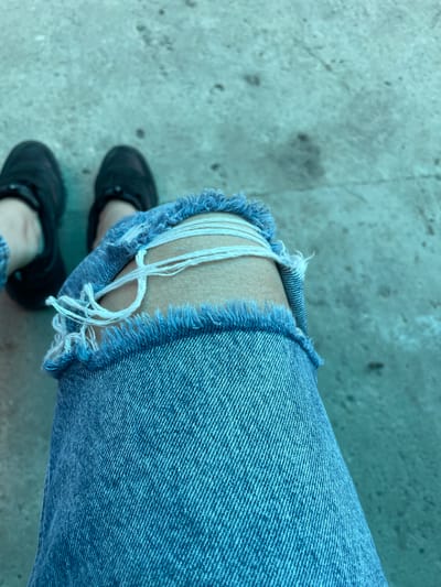 The image depicts a person wearing ripped blue jeans with black shoes standing on a concrete surface. The rips in the jeans expose the skin of the leg and strands of white fabric. The concrete surface has a faded appearance and no specific details indicate the location is Olginka, Russia. No specific actions or interactions are evident.