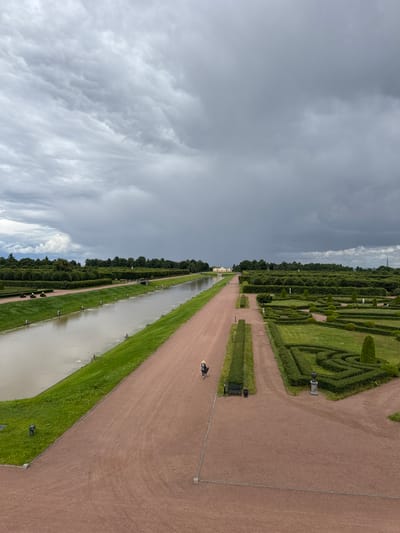 The image shows a wide aerial view of the Strelna, Russia gardens, specifically the Constantine Palace grounds. The scene is dominated by a long, rectangular canal on the left, bordered by a strip of vibrant green grass and a pink gravel pathway. A single figure, a person wearing a hat and dark clothing, is walking along this pathway. To the right of the pathway, a meticulously manicured hedge maze and formal garden areas are visible. These gardens feature geometric patterns of trimmed hedges and scattered sculptures. In the background, a long, yellow building, likely part of the palace complex, is visible across the canal, with dense green trees framing the horizon. The sky above is a dramatic and overcast gray, with dark, heavy clouds suggesting an impending storm. The overall impression is one of grand, formal landscaping under a moody sky.