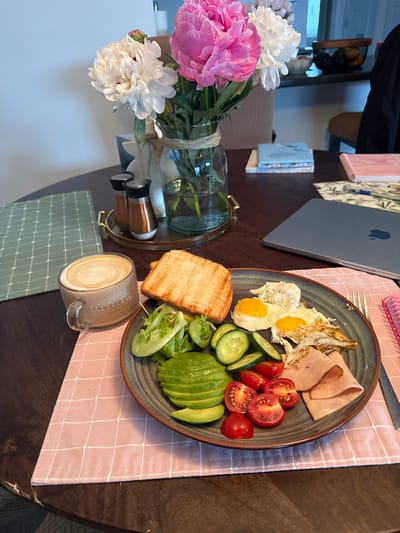 A top-down, slightly angled shot shows a breakfast spread on a dark wooden table, likely in a home dining area. The meal is presented on a grey, speckled plate and consists of two fried eggs, sliced ham, halved cherry tomatoes, cucumber slices, sliced avocado, and a small pile of mixed greens. To the side of the plate is a piece of toasted bread. A glass mug filled with coffee and latte art sits to the lower right of the plate, on a green checkered placemat. A pink checkered placemat lies beneath the plate and coffee cup.
To the left of the table setting, a bouquet of pink and white peonies is arranged in a clear glass vase. A brass tray with two small spice shakers is behind the vase. In the background, a glimpse of a laptop with an Apple logo is visible, along with several small notebooks. The overall impression is that of a leisurely breakfast or brunch. The lighting suggests it could be daytime, possibly morning or early afternoon. There are no people visible in the image.
