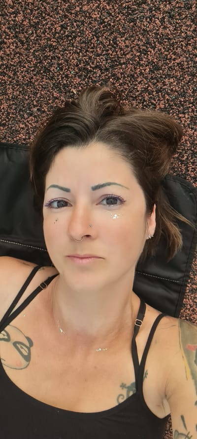 A woman lies on a black surface, likely a cushion or mat, with what appears to be a dark brown and red artificial turf background. She has dark hair, fair skin, and both a nose and nostril piercing on the left side of her nose. Some form of glitter or shimmering material is present around her left eye and on her chest. She is wearing a black tank top with thin straps. Her tattoos are visible on her arms and chest.

It is not possible to confirm the location in the city of Kiryat Gat, Israel, or to comment on actions, interactions, or the precise environment without further information.
