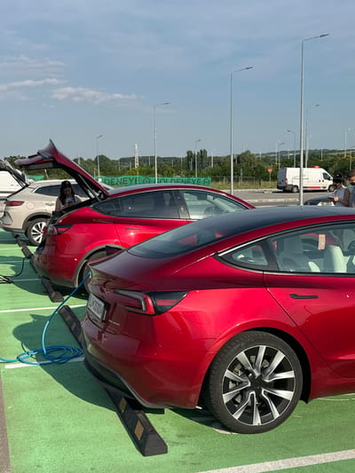 This detailed description outlines a scene at an electric vehicle charging station located at the Kapitan Andreevo border crossing in Bulgaria, evidenced by the "DENEYE GOLDENEYE" signage visible in the background.
The image is captured outdoors under a clear blue sky with scattered cumulus clouds, illuminated by direct sunlight. The ground surface consists of a vibrant green material, likely painted asphalt or artificial turf, marked with white lines defining individual parking bays. Each bay is equipped with black and yellow striped parking stoppers.
Three vehicles are prominent in the midground and foreground. The central vehicle is a red Tesla Model 3, parked facing away from the viewer with its trunk open. A coiled blue electric charging cable is connected to its rear-left charging port, extending towards the left side of the frame where the charging apparatus is partially obscured. The license plate "L4-OS1004" is visible on this Tesla. A woman with dark hair and glasses, wearing a striped top, stands near the open trunk, looking down at a mobile phone in her hands. Directly behind this Tesla, a light beige SUV with a multi-spoke wheel design is also parked with its trunk open.
In the foreground, on the right side of the image, the rear section of another red Tesla Model 3 is visible. This vehicle displays a rear wheel with black and silver multi-spoke rims and features light-colored interior seats, visible through the rear window.
The background reveals a landscape of rolling hills covered in trees. Numerous tall, slender streetlights are interspersed across the area, alongside visible power lines and utility poles. A large green sign prominently displays "DENEYE" and "GOLDENEYE" in white lettering. On a road beyond the charging station, a white delivery van with red rear markings is visible. A red circular stop sign is partially visible near the road. On the far right edge of the frame, two individuals are partially visible, appearing to be standing near a dark vehicle. One person, wearing a white top and a face mask, faces left towards the scene.