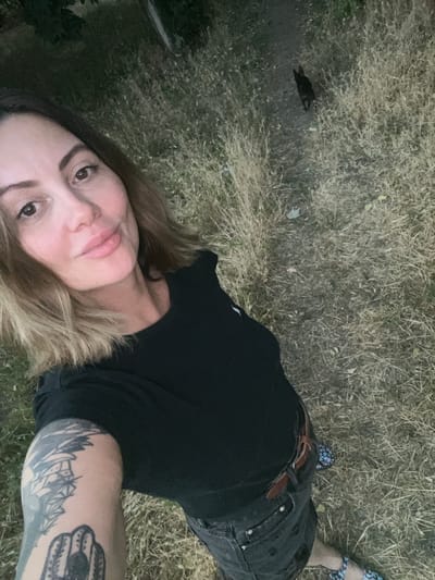 In Voznesensk, Ukraine, a woman with blonde hair and a tattoo on her left arm is taking a selfie outdoors. She is wearing a black t-shirt and dark jeans. Her feet are clad in colorful sandals. A black cat is visible in the background, walking on a dirt path through dry grass. The environment appears to be a rural or overgrown area, possibly a park or a field on the outskirts of the city. The lighting suggests it might be late afternoon or early evening.