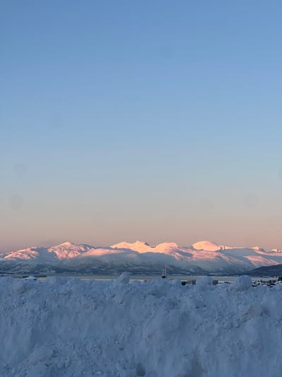 The image displays a serene winter landscape, likely taken during either sunrise or sunset, given the warm, golden light illuminating the mountain peaks. The perspective is from ground level, looking towards a distant mountain range.

In the foreground, on the right side of the image, there is a large, irregularly shaped mound of rough, white snow, suggesting plowed snowdrifts or accumulated snow. The texture of this snow is uneven and appears quite deep.

Immediately behind this snowdrift, stretching across the middle ground, is a flat, dark grey or asphalt-colored surface, possibly a road, runway, or cleared area, which appears to lead towards the distant mountains. There are a few small, indistinct structures or vehicles visible along this flat expanse.

Dominating the midground and background is a majestic range of snow-capped mountains. The peaks are brightly illuminated by the low-angle sunlight, casting a warm, almost pinkish-orange glow on the snow. The slopes facing away from the sun are in shadow, appearing in cooler shades of blue and grey. The mountains extend horizontally across a significant portion of the image, fading into the distance.

The sky occupies the upper two-thirds of the image. It presents a gentle gradient, transitioning from a clear, light blue at the top and left to a warmer, pale yellow or peach hue closer to the horizon, blending with the light hitting the mountains. There are no visible clouds.

The overall impression is one of cold, clear weather, with the low sun creating dramatic lighting on the snowy terrain. There is no discernible text in the image.