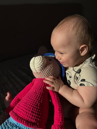 In Yanino-1, Russia, a young baby is seen in a dimly lit room, possibly a bedroom. The baby, with light blonde hair, is looking down at a large, crocheted stuffed animal. The toy has a white and beige head with a blue and brown knitted hat, and a vibrant pink body with blue knitted legs. The baby is wearing a light-colored t-shirt with a black pattern of faces. The scene is illuminated by a strong beam of light coming from the left, casting deep shadows across the dark, textured surface the baby and toy are resting on. The baby's hand gently touches the stuffed animal, suggesting a moment of quiet interaction.