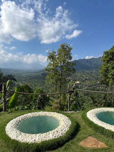 The image depicts an outdoor scene in Sidemen, Indonesia, featuring two circular pools filled with turquoise water, set within a grassy area. The pools are bordered by a ring of smooth, white stones. In the foreground, lush green grass surrounds the pools, and a rough-hewn stone path leads between them. Beyond the pools, a wooden fence with rope accents separates the foreground from the background. The mid-ground reveals a verdant landscape with dense tropical vegetation, including palm trees and banana leaves, leading to a hillside dotted with traditional Balinese buildings and thatched roofs. In the distance, misty mountains rise against a bright blue sky dotted with fluffy white clouds. The overall impression is one of a serene and picturesque retreat.