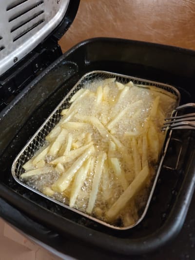 The image shows a close-up of French fries being deep-fried in a metal basket within a black electric deep fryer. The fries are golden yellow and bubbling vigorously in hot oil. The lid of the deep fryer is open, revealing its metallic interior with ventilation slits. The fryer is placed on a surface with a light brown, textured appearance, possibly a countertop or table. There is a metal fork visible on the right side of the image, resting on the edge of the fryer basket. The scene is domestic, focusing on the cooking process. The image does not contain any elements that can be used to determine its location in the city of Bor, Russia.