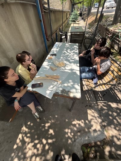 In Badiauri, Georgia, four individuals are gathered around a white table outdoors. Two young males are seated on a wooden bench to the right, both consuming food. One young female, wearing glasses and a yellow shirt, is seated at the table, also eating. Another young female, in a black shirt, is seated to the left of the table and appears to be watching the others. Bread is visible on the table, along with a mobile phone and a plastic bag. The environment suggests a casual outdoor dining area with a concrete floor and dappled sunlight filtering through tree leaves. A wall borders the scene on the left, and a street with vehicles is visible in the background.