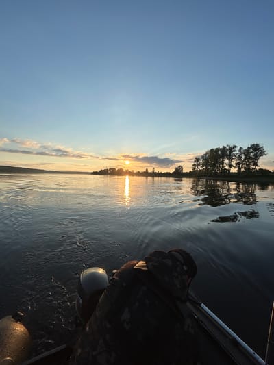 This media file depicts a serene scene of a person in a boat on a body of water at sunset. The sky is a clear, pale blue, transitioning to warm hues of orange and yellow near the horizon, indicating either sunrise or sunset. Wispy clouds are scattered across the sky, tinged with pink and gold from the setting sun.
The water is calm, with gentle ripples reflecting the colors of the sky and the silhouette of trees along the distant shoreline. These trees are densely packed, forming a dark, irregular line against the brighter sky. A bright band of sunlight shimmers across the water, creating a path of light from the horizon towards the foreground.
In the foreground, the back of a person wearing a camouflage jacket and a cap is visible. The person appears to be seated or standing in a boat, as part of a metallic railing is visible to the right. A rounded, light-colored object, possibly a part of the boat's equipment or a buoy, is also visible near the person. The overall mood is peaceful and contemplative, suggesting a moment of quiet enjoyment of nature. The presence of the boat and the water implies a recreational activity like fishing or simply cruising. The time of day is clearly around sunset, given the quality of light and sky colors. No text is visible in the media file.