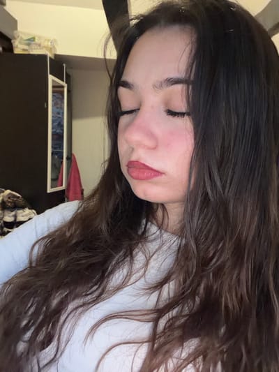 A woman with long, wavy dark hair and fair skin is featured. Her eyes are closed, and she has a slight, natural-looking makeup on, particularly around her lips and cheeks. The background includes a dark-colored dresser or cabinet with visible items on top, some covered by a red fabric. The woman is wearing a white top. The image is taken from a slightly high angle and rotated, giving a casual, almost candid feel.
