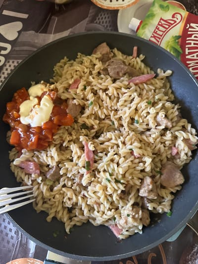 A close-up overhead shot shows a serving of fusilli pasta with meat and sauce in a dark gray frying pan. The pasta is mixed with chunks of meat, possibly beef or pork, and pieces of what appears to be salami or ham. Chopped green onions are also visible throughout the dish. On the left side of the pan, a dollop of white sauce, likely mayonnaise or sour cream, sits atop a portion of red sauce with visible chunks of vegetables. A silver fork with three tines is resting on the rim of the pan on the lower left side. In the background, a tube of tomato paste with Russian labeling is partially visible. The scene is set on a patterned tablecloth. There is no discernible indication of the city of Gorno-Altaysk, Russia.