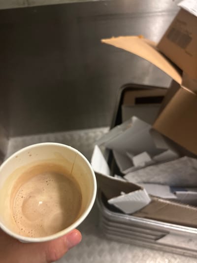 A hand holds a white paper cup filled with a frothy, light brown liquid, likely coffee, positioned over a stainless steel sink. The cup is about half full and the surface of the liquid is covered in a thin layer of foam with small bubbles. In the background, a cardboard box is tilted open, revealing a jumble of packing materials and possibly other discarded items. The scene appears to be in an indoor setting, with the stainless steel sink suggesting a kitchen or work area. The lighting is somewhat dim, casting shadows. The overall impression is one of a casual, everyday moment, possibly a break in a workday or a quick coffee before starting a task. There is no visible text in the image.