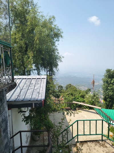 The image depicts a view from a raised vantage point in Dharamshala, India, overlooking a valley. In the foreground, a section of a white building with a corrugated metal roof is visible on the left. A green metal railing lines a concrete staircase that descends from the viewer's perspective. Lush green trees with sparse foliage dominate the midground and left side of the image. Beyond the trees, a hazy valley unfolds, dotted with buildings and vegetation, suggesting a populated area. The sky is a clear light blue with a single, small white cloud. Prayer flags are strung across a railing in the midground, adding a cultural element to the scene. The overall impression is one of a serene, elevated location offering a panoramic view of the surrounding landscape.