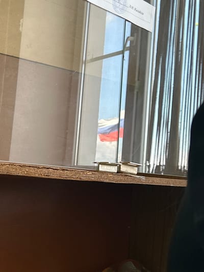 The image shows a view through a glass case. Reflected in the glass is a Russian flag. To the left is a piece of paper with Russian text. The case is made of wood. The location may be in Dmitrievskaya, Russia.