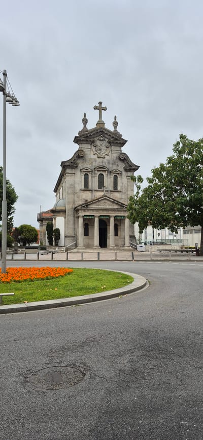 The image shows the Igreja de Nossa Senhora da Conceição, a church located in Póvoa de Varzim, Portugal. The church has a neoclassical facade with a large stone cross at the top. In front of the church, there is a circular garden with orange flowers and green grass, bordered by a gray concrete curb. A paved road with a manhole cover in the foreground curves around the garden. A street lamp is visible to the left of the church. A tree with green foliage stands to the right of the church. The sky is overcast. There are no people visible in the image.