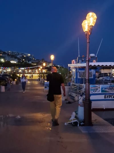 The image depicts a waterfront scene at dusk in Sveti Vlas, Bulgaria. A man in a black t-shirt and yellow pants walks away from the camera on a paved walkway. To his right, a boat with blue and white trim and the Cyrillic text "БУРГАС" on its side is docked. Behind the boat, other vessels are visible. In the background, buildings are illuminated on a hillside, suggesting a town or resort area. Decorative lights strung along a building facade add to the ambiance. A street lamp with two glowing diamond-shaped fixtures illuminates the foreground. The ground appears wet in places, reflecting the ambient light.