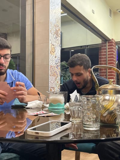 Two young adult males are seated at a black, reflective table within a cafe or restaurant in the city of Homs, Syria. The male on the left, wearing glasses and a light blue polo shirt, is holding a fan of reddish-brown playing cards. His gaze is directed downwards. The male on the right, wearing a dark top, has an open-mouthed expression, suggesting a reaction or vocalization.
On the table are various items: a traditional ornate metallic teapot, multiple empty clear glasses (some textured), a glass containing a light-colored beverage in a mint-green silicone holder with a spoon, several playing cards laid flat, crumpled white napkins, and a white-cased smartphone.
The background features a decorative square pillar adorned with colorful mosaic-like tiles in geometric and floral patterns, large glass panels reflecting interior lighting and showing glimpses of a dark exterior, and a reddish-brown brick column. A small white surveillance camera is mounted high on a wall to the right. Green foliage is faintly visible behind the right male. A teal-colored upholstered chair or bench is partially visible under the table.