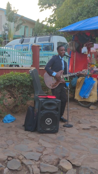 A musician plays an electric guitar while standing on a stone path. He wears a dark suit with a light-colored shirt. A microphone is positioned in front of him, and a large speaker sits on the ground next to his open guitar case. A colorful market stall and a white vehicle are visible in the background, partially obscured by a blue tarp. Vegetation borders the scene, and the lighting suggests it is either morning or late afternoon.