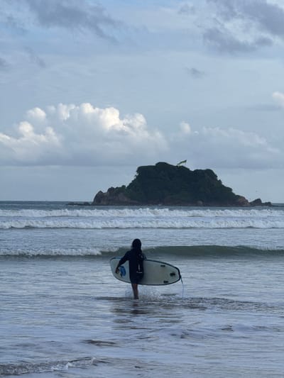 An overcast day at Weligama beach in Sri Lanka, where a lone surfer, dressed in a black wetsuit, is wading into the ocean with a light-colored surfboard. The surfer's back is to the camera, and they are walking away from the shore towards the sea. The surfboard is positioned under their right arm, and a blue leash is visible trailing from it. In the background, several layers of waves roll towards the beach. Further out, a lush green island with a rocky outcrop and a flag flying from its summit stands in the sea. The sky is filled with dramatic, cloudy formations.