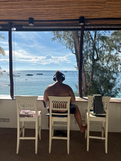 An individual is seated with their back to the camera, looking out over the ocean from an elevated open-air establishment in Koh Tao, Thailand. The person is bare-chested, wearing dark patterned shorts, and has dark hair gathered at the back of their head, wearing black over-ear headphones. They occupy a white wooden high-backed chair featuring decorative cutouts on its backrest. To the left, an identical unoccupied chair holds a red and white striped cloth on its seat. To the right, another identical unoccupied chair has a dark geometric-patterned cloth draped over its backrest, and a plate containing a dark, circular object on its seat. The structure's ceiling is constructed from bamboo slats, supported by dark vertical beams. A white concrete ledge serves as a railing or counter, separating the interior from the expansive view of the calm blue-green sea under a partly cloudy sky. Several boats, including smaller local vessels and at least two white motor yachts, are visible in the water. Lush green foliage, including palm fronds on the far left and dense trees on the right, frames the marine panorama.