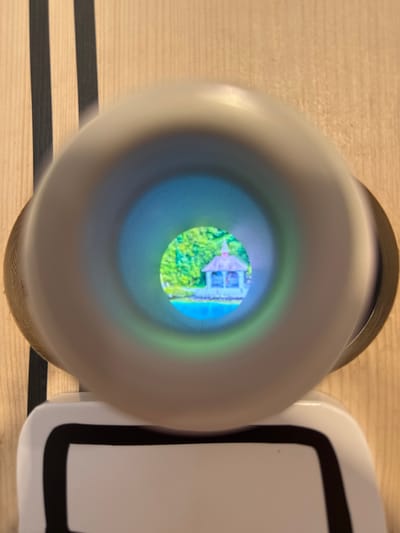 This image captures a close-up view of a peephole, through which a miniature scene of a gazebo beside a body of water is visible. The gazebo, with a pointed roof and visible pillars, appears to be located on a small island or peninsula. Lush green foliage surrounds the water, suggesting a natural, possibly park-like setting. The scene within the peephole is brightly lit and colorful. The outer elements of the image show the textured wooden surface of the object containing the peephole, with two vertical black stripes on the left side. A portion of a white object with black trim is visible at the bottom of the frame. This image is taken at the Uri Museum in Schattendorf, Switzerland, specifically in the "Museum im Ballade" exhibition, which features a diverse collection of objects, including those that offer unique perspectives or visual experiences.