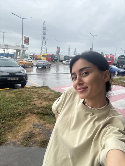 The image depicts a street scene with a woman in the foreground, taking a selfie. She has dark hair and is wearing a light-colored t-shirt and a pearl necklace. Behind her, a parking lot is filled with various vehicles, including cars and trucks. The ground is wet, indicating recent rain, and there are visible puddles and reflections. In the background, there are utility poles with high-voltage wires and a large tower-like structure. Some buildings and signs are also visible, suggesting a commercial or industrial area. The overall environment appears to be outdoors, possibly a rest stop or truck stop, given the presence of numerous large trucks. The sky is overcast, contributing to a muted lighting. The location is identified as being in the city of Argveta, Georgia.