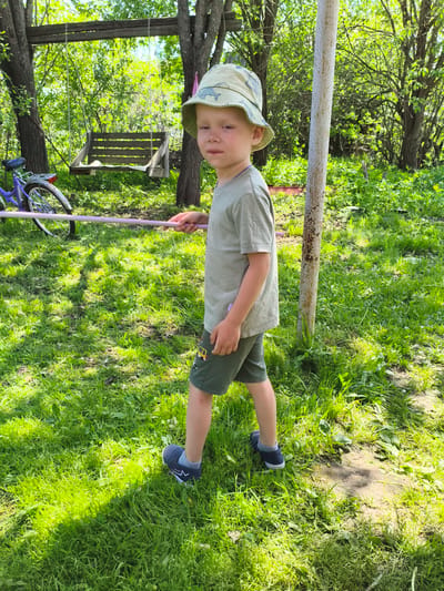 A young boy stands in a grassy yard in Chernaya, Russia, holding a purple stick. He wears a bucket hat, a short-sleeved t-shirt, shorts with cartoon car details, and blue shoes. A rusted metal pole is positioned beside him, and in the background, there is a bicycle and a wooden swing hanging from trees. The environment is filled with lush greenery.