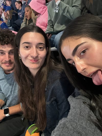 The image depicts three individuals in the foreground, seated in stadium-style spectator seating, likely at MetLife Stadium in East Rutherford, New Jersey, United States.

In the center foreground, a young adult female with long dark brown hair and a dark denim jacket over a grey top looks directly at the camera with a subtle smile and pursed lips. To her left, a young adult male with curly dark hair, light facial hair, and a grey t-shirt smiles towards the camera. He wears a silver chain and a white smart watch on his left wrist. To the right of the central female, a second young adult female with long dark hair, wearing a grey knitted sweater, partially faces the camera with her tongue extended. An orange and green object is visible at the bottom of the frame, below the central female.

In the blurred background, additional spectators are seated. One individual wears a sparkling pink hooded sweatshirt, and another wears an olive-green hooded sweatshirt with a white tag attached. A person further in the background wears a blue cap and a shirt with "NEW YORK GIANTS" visible text, confirming a sports event context. The scene is illuminated by artificial lighting, consistent with an indoor or evening event.