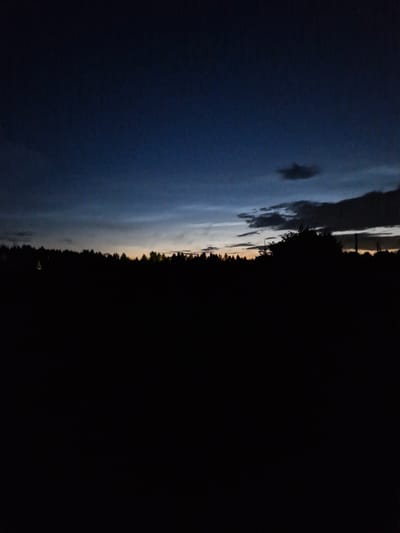 The image captures a twilight scene over a silhouetted landscape. The sky is a deep, gradient blue, transitioning to lighter shades near the horizon where faint hints of orange and yellow suggest the last vestiges of sunlight. Several clouds are visible, some dark and heavy, others wispy and lighter. Below the sky, a dense line of trees forms a dark silhouette, suggesting a forest or a wooded area. The foreground is almost entirely black, indicating a very dark or unlit area, possibly a field or open ground. There are no visible people or specific structures that can be identified with certainty due to the low light conditions. The overall impression is one of a peaceful, natural environment at dusk or dawn. The location is identified as Sorochkino, Russia.