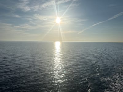The image displays a vast expanse of calm ocean under a bright, partially cloudy sky. The sun is prominently featured in the upper center of the frame, casting a strong glare and creating a shimmering reflection on the water's surface. The horizon line is clearly defined, separating the deep blue of the sea from the pale blue and white of the sky. Wispy clouds stretch across the sky, some forming contrails. The water exhibits gentle ripples, with a visible wake trailing off to the right side of the frame, suggesting movement. The scene is devoid of any human presence or man-made structures, presenting a natural and serene seascape. The location is stated as Unknown, Sweden.
