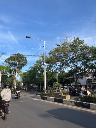 The image depicts a street scene in Langsa, Indonesia, under a clear blue sky with scattered white clouds. The street is paved with asphalt and has a yellow dividing line. On the right side of the road, there is a concrete median with black and white markings, lined with planters containing small trees and bushes. Beyond the median, a sidewalk is visible with several individuals on motorcycles. A prominent streetlight with two lamps stands in the middle of the scene, along with power lines. Trees with dense green foliage are present along the street, casting shadows on the road. Buildings and shops are visible in the background, including a sign that reads "Bank Aceh". Several people are riding motorcycles and one individual is standing near a motorcycle. The overall atmosphere suggests a typical day in a city with a tropical climate.