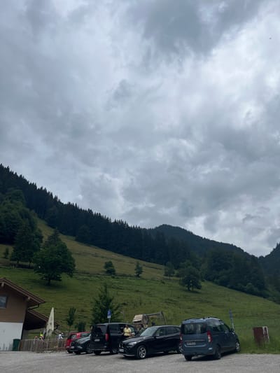 The image shows a parking lot in front of a building with a wooden facade, likely a restaurant or guesthouse, nestled at the base of a grassy, tree-covered hill under a cloudy sky. The location is identified as Ettal, Germany. In the parking lot, several vehicles are parked: a red car, a black car, a black van, and a blue van. A few people are visible near the building and by the parked cars. The hill behind the parking lot is covered in green grass and dense coniferous trees, with a lighter, possibly wooded area higher up. The sky is overcast with heavy gray clouds.