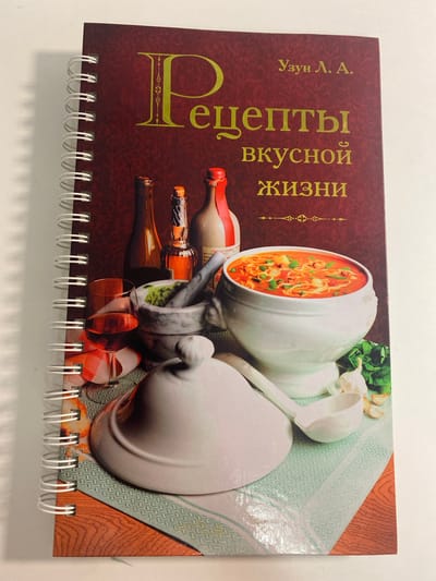 The image shows the front cover of a spiral-bound cookbook. The cover is a dark red color with gold text. The title, "Рецепты вкусной жизни," which translates to "Recipes for a Delicious Life," is prominently displayed in large, ornate gold lettering. Above the title, in smaller gold text, is the name "Узун Л. А." The lower half of the cover features a still life arrangement of culinary items. In the center, a large white tureen overflows with a vibrant red soup, likely a tomato-based dish with pasta and other ingredients visible. To the left of the tureen is a mortar and pestle filled with green ingredients, possibly herbs or spices. Behind the mortar and pestle, two bottles of liquid and a glass of red wine are arranged. A white ladle rests on the table beside the tureen. In the lower right corner, a head of garlic and a few cloves are scattered, along with some green herbs, likely basil. The background of the cover appears to be a patterned tablecloth in shades of teal and white, with a section of wood grain texture at the bottom. The spiral binding is white. The cookbook is displayed on a white surface, possibly a table or a wall, with a shadow cast to the left. The location is not mentioned in the image.