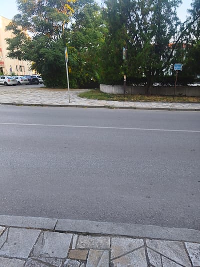 A street scene in Harmanli, Bulgaria, shows parked cars, trees, and a sidewalk with a road. The cars are white and dark-colored, with at least three visible. The trees are dense and green, providing shade. A concrete wall separates the sidewalk from the trees. A street sign is visible on the right side of the image. The road is paved and has a white line marking its center. The sidewalk is made of stone tiles. The overall lighting suggests it is daytime.