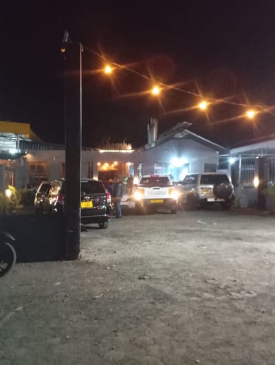 The image shows a parking lot at night in Arusha, Tanzania. There are five vehicles parked in the lot: a black SUV, a silver SUV, a white pickup truck, and two other cars. The vehicles are parked facing towards the camera.  The white pickup truck has the word "ISUZU" written on the back.  The black SUV has a license plate that is visible in the image.  There are two people walking in the background, both wearing dark clothing, near the white pickup truck. There is a sign above the white pickup truck with the word "Shambe" on it.  There are several string lights strung across the top of the parking lot.  There is a street light illuminating the area in the background. The parking lot is made of gravel.  There is vegetation in the background of the image to the right.  The image is captured from a low angle looking up towards the parking lot.  The location of the parking lot is a business district in Arusha, Tanzania.
