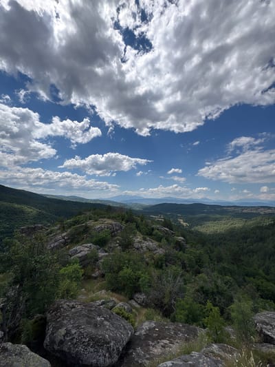 The image is a high-angle shot of a mountainous, forested landscape on a partly cloudy day. In the foreground, large, weathered grey boulders are scattered across a rocky outcrop covered with sparse green vegetation and dry grass. Beyond this, a dense, dark green forest covers the slopes of rolling hills, which recede into the distance. A valley with scattered trees and fields is visible between the hills. Further in the background, layers of lighter green and blue mountains fade into the hazy sky. The sky itself is a bright blue, populated with a dynamic mix of white and grey clouds, some of which are illuminated by sunlight. The overall impression is a vast, natural landscape, likely within the vicinity of Dolno Drianovo, Bulgaria, given its hilly terrain and forested expanses. No people or animals are visible in the scene.