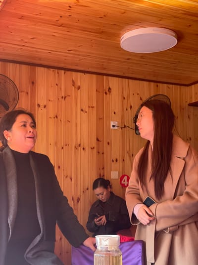 Inside a wood-paneled room, three women are present. One wears a tan coat and glasses, another a dark jacket, and the third a dark coat and light gray scarf. A round, flat light fixture is affixed to the wall. Electrical outlets and a red circle with the number "4" are also visible on the wall. A fan sits on the floor.