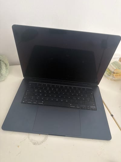 The image displays a dark gray MacBook laptop, open and placed on a light-colored, possibly textured surface. The laptop is angled so that the keyboard and trackpad are clearly visible, along with a portion of the screen. The screen is dark, suggesting it is either off or displaying a dark image. The keyboard has a German layout, indicated by the presence of keys such as "Ü," "Ö," and "Ä." The trackpad is large and centrally located below the keyboard.
In the background, to the upper left of the laptop, a clear plastic container with some food items, possibly snacks or pastries, is partially visible. The surface the laptop rests on appears to be a table or desk, and there are some faint smudges and crumbs scattered on it, hinting at a casual or recently used setting. A light-colored, textured wall forms the background on the left side of the image. The lighting suggests it might be daytime, but the absence of windows or clear environmental cues makes it difficult to determine the exact time of day or weather conditions. The scene is static, with no activity taking place. The overall impression is one of a personal workspace or a moment of rest during work or study. No specific emotions are conveyed, and the focus is on the object itself and its immediate surroundings in Saarbrücken, Germany.