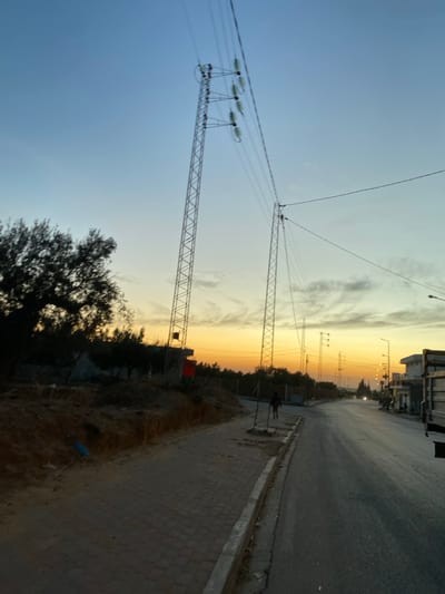 An outdoor, landscape-style photo captures a street scene at dusk or dawn. Power lines and lattice towers dominate the left side, reaching towards a sky transitioning from blue to orange. Buildings are visible in the distance on the right side. A road and a sidewalk run along the right side of the frame. A leafy tree and some vegetation are on the right side, closer to the viewer.