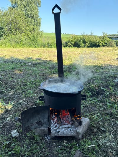 In the village of Permiaki, Russia, a large black cauldron filled with boiling water sits atop a makeshift fire pit. The fire pit is constructed from metal and is fueled by burning wood, with visible flames and embers. Smoke billows from the cauldron, indicating active cooking. The scene is set outdoors, with green grass and vegetation surrounding the fire pit. In the background, a field and trees are visible under a clear blue sky. The overall impression is one of outdoor cooking, likely a traditional meal preparation.
