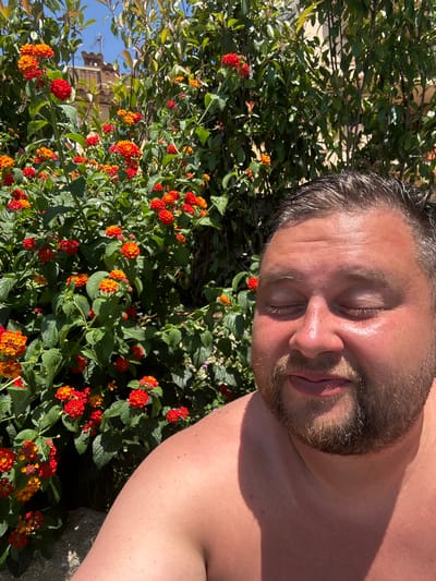 An outdoor, close-up, eye-level, medium shot captures a man with his eyes closed, his face tilted upwards towards the sun, basking in the light. He has brown hair, a beard, and appears to be shirtless. His skin is flushed, indicating exposure to the sun. Behind him, a dense bush of vibrant orange and red Lantana flowers is in full bloom. The background also shows glimpses of green foliage and a blurred architectural element with a terracotta roof and a small turret, suggesting an urban or semi-urban setting. The scene is likely Sant Feliu de Guíxols, Spain, given the distinctive architecture and flora. The overall mood is one of relaxed enjoyment of a sunny day.