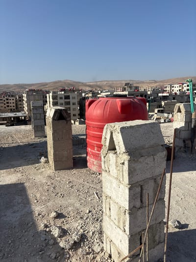 The image depicts an elevated view of a cityscape under a clear blue sky in Mu'adammiyat ash-Sham, Syria. In the foreground, a dusty, gravel-covered rooftop features several unrendered concrete block structures, one of which has a distinct triangular top. A large, red, cylindrical plastic water tank stands prominently in the mid-ground. Near the concrete structures, several rusty metal rebar rods protrude vertically from the ground. The urban environment extends into the background, showcasing numerous multi-story residential buildings, many with visible water tanks on their rooftops and some appearing unfinished or under construction. Distant, arid hills frame the horizon, with a faint plume of smoke or haze visible on the left side. No individuals are present in the scene. The lighting suggests bright daylight, casting strong shadows from the foreground objects.
