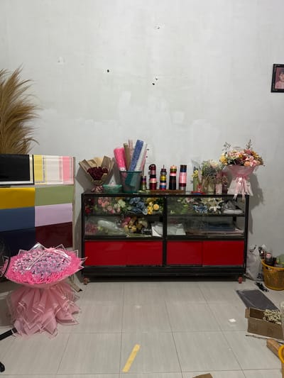 The image depicts a flower shop in Gampongasanlhoksukon Barat, Indonesia. In the center of the image is a display cabinet with a black frame and red drawers. The top shelf of the cabinet holds various floral arrangements and supplies, including a bouquet of red roses, colorful artificial flowers, and rolls of decorative paper and ribbon. The lower shelves are filled with more artificial flowers and floral arrangement materials. To the left of the cabinet, stacks of colorful wrapping paper are neatly arranged. In the foreground, a large bouquet wrapped in pink tulle and ribbon is prominently displayed. The background is a plain, light-colored wall, with a framed photograph of a child hanging on the right side. Various supplies, including a yellow basket filled with items and several plastic bags, are visible on the floor to the right of the cabinet.