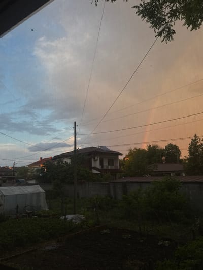 The image depicts a residential scene in Elhovo, Bulgaria. A two-story house with a red roof and solar panels is visible, surrounded by greenery and a concrete fence. Utility poles and power lines dominate the sky, which transitions from blue with clouds to a light orange hue, possibly indicating sunrise or sunset. A portion of a mesh screen is visible in the foreground. Adjacent buildings and vegetation suggest an urban or suburban setting. A parked or covered vehicle can be seen on the right side. No people are visible in the image.
