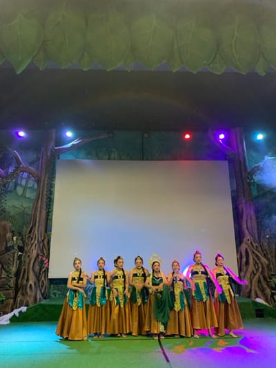 Nine women are standing on a stage in front of a white screen in the Jatim Park 2, Batu, Indonesia.  All women are wearing long gold dresses with green and blue designs and matching headbands. The woman in the middle of the line is wearing a green shawl. The stage has green padding and there are spotlights shining down on the women. The backdrop is a large screen with a jungle scene.  The stage is illuminated by purple and pink spotlights. The ceiling is decorated with fake green leaves.  The location is Jatim Park 2, Batu, Indonesia (latitude: -7.885, longitude: 112.524).