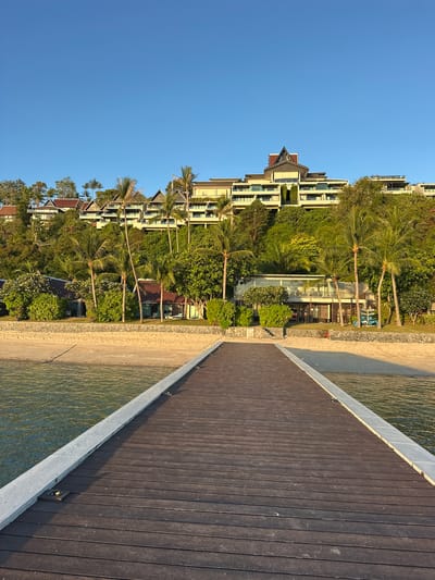 A wooden pier extends from the foreground into a body of calm, clear water with a sandy beach to the right. Beyond the beach, a resort complex is visible, built into a lush, green hillside under a clear blue sky. The resort features multiple levels of white buildings with dark roofs, interspersed with palm trees and other tropical foliage. A few smaller buildings with tiled roofs are situated closer to the beach, with some lounge chairs visible on the sand. The overall scene depicts a serene tropical resort environment.