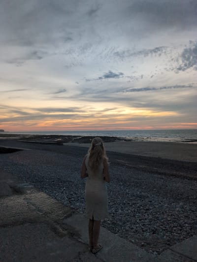 The image depicts a woman standing on a pebbled walkway overlooking the sea at dusk. The sky is filled with clouds, painted with hues of orange, yellow, and grey, indicating either sunrise or sunset. The sea is calm, with a visible coastline in the distance. The woman is facing away from the camera, her blonde hair tied up with a light-colored ribbon. She is wearing a light-colored dress and patterned shoes. The walkway is made of concrete and gravel, with patches of water reflecting the sky on the concrete section. The overall scene suggests a tranquil moment by the sea. This location appears to be the seaside promenade in Veules-les-Roses, France, a picturesque village known for its beaches and charming atmosphere.