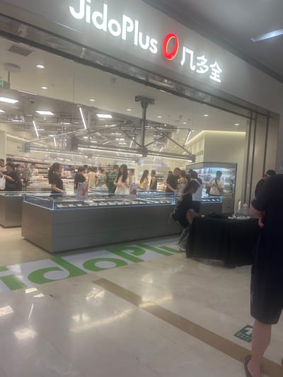 This is a medium shot of the interior of a brightly lit, modern supermarket. The store features a large, illuminated sign above the entrance that reads "JidoPlus 凡多全". Inside, various display cases are visible, stocked with items, and several shoppers are browsing. The floor is polished marble with a green directional arrow graphic on it. In the foreground, a black fabric-covered table is set up, with a few white cube-shaped objects on top. A person in a dark t-shirt and shorts is standing near the table, partially obscuring it. The overall impression is a busy retail environment. This image was taken in Liandu District, China.
